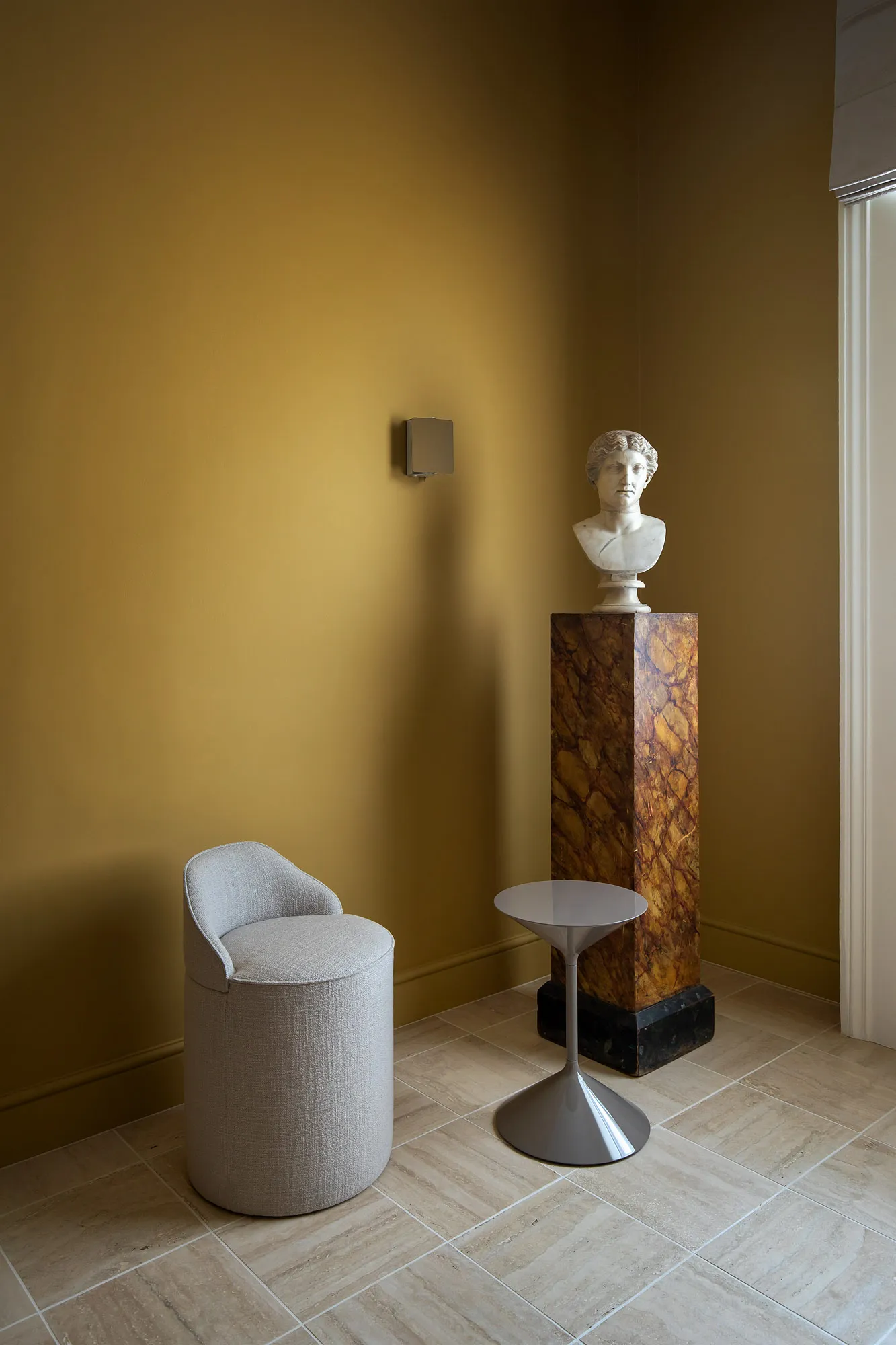 Quiet ochre room with a white chair, small table and sculptural bust